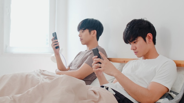 Casal gay asiático usando telefone celular em casa. jovens asiáticos lgbtq + homens felizes relaxam descansar juntos depois de acordar, verifique as mídias sociais, deitada na cama no quarto em casa de manhã.