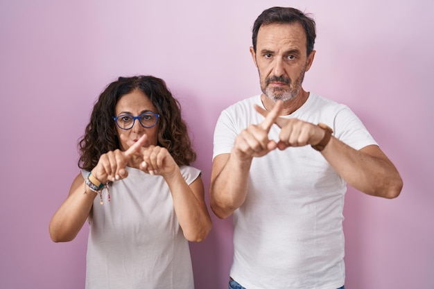 Casal hispânico de meia-idade juntos sobre fundo rosa expressão de rejeição cruzando os dedos fazendo sinal negativo