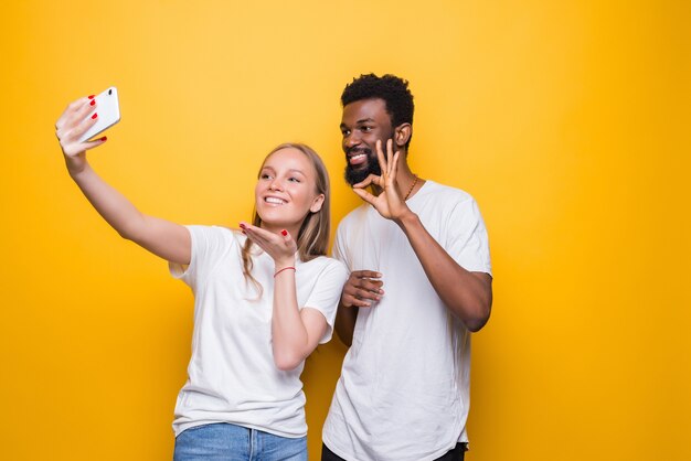 Casal interracional alegre fazendo autorretrato juntos, olhando para a frente e sorrindo, posando sobre a parede amarela