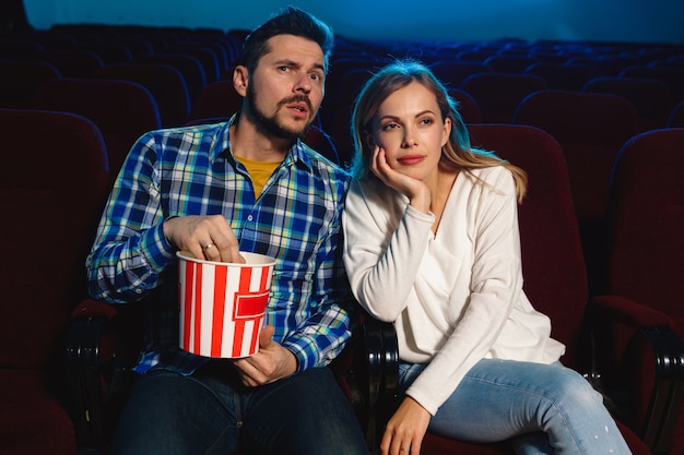 Casal jovem atraente caucasiano assistindo a um filme no cinema, casa ou cinema. pareça expressivo, surpreso e emocionado. sentado sozinho e se divertindo. relação, amor, família, tempo de fim de semana.