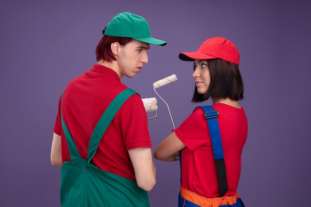 Casal jovem cara sério garota confiante com uniforme de trabalhador da construção civil e boné em pé atrás de vista segurando o rolo de pintura, olhando um para o outro isolado na parede roxa