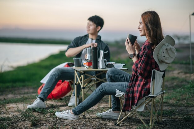 Casal jovem e alegre de mochileiros sentado em frente à barraca na floresta com um conjunto de café e fazendo um moedor de café fresco durante o acampamento nas férias de verão. Foco seletivo