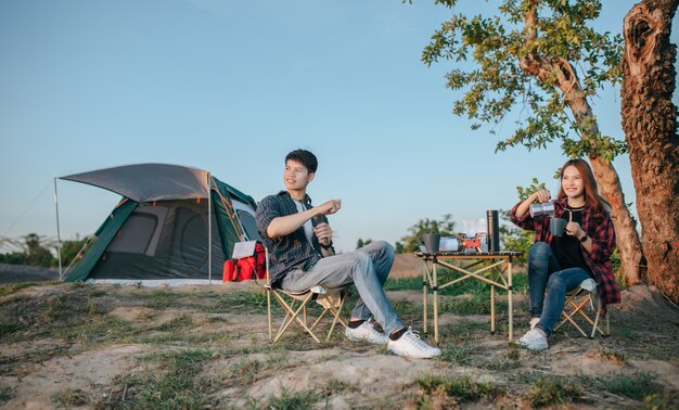 Casal jovem e alegre de mochileiros sentado em frente a uma barraca na floresta com um conjunto de café e fazendo um moedor de café fresco durante o acampamento nas férias de verão