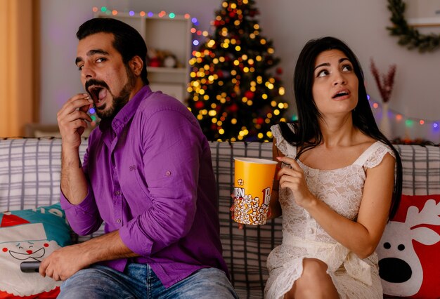 Casal jovem e bonito feliz alegre homem e mulher perplexa com balde de pipoca assistindo tv juntos em quarto decorado com árvore de natal na parede