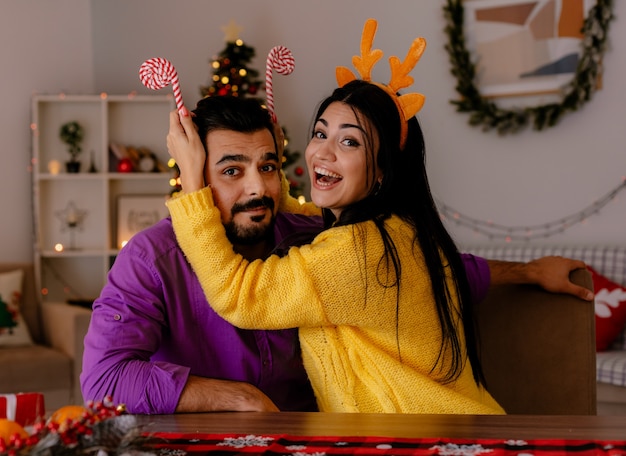 Casal jovem e bonito, homem e mulher com bastões de doces, se divertindo juntos, felizes e apaixonados em um quarto decorado de natal com uma árvore de natal ao fundo