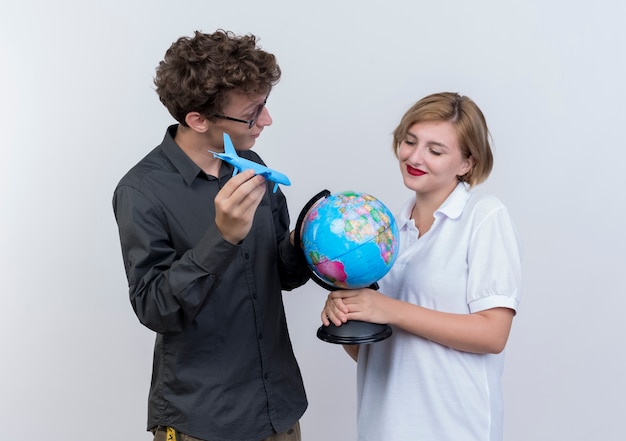 Foto grátis casal jovem e feliz de turistas, homem e mulher segurando um avião de brinquedo globo e pé juntos sobre uma parede branca