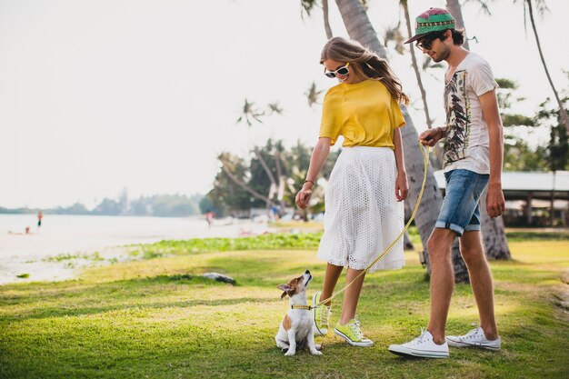 Casal jovem elegante e hippie apaixonado andando e brincando com um cachorro em uma praia tropical