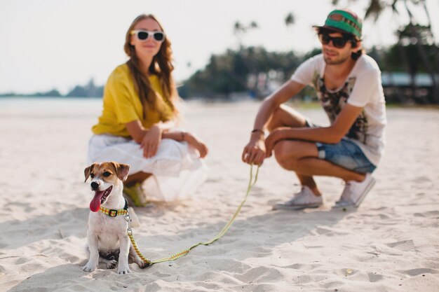 Casal jovem elegante e hippie apaixonado andando e brincando com um cachorro em uma praia tropical