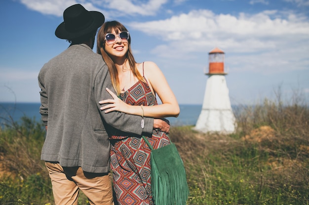 Casal jovem hippie estilo indie apaixonado caminhando pelo campo, farol no fundo