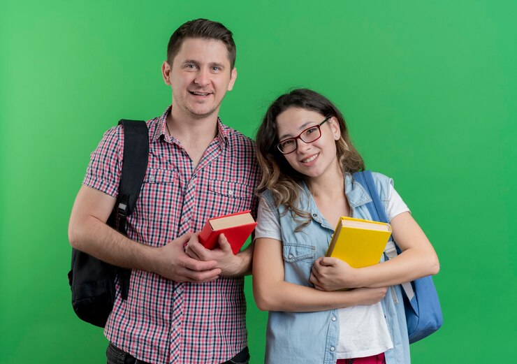 Foto grátis casal jovem, homem e mulher com roupas casuais e mochilas segurando livros, sorrindo amigável em pé sobre a parede verde