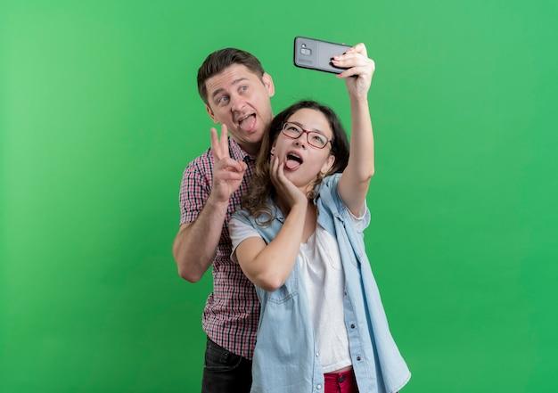 Casal jovem, homem e mulher com roupas casuais, juntos fazendo selfie, feliz no amor, se divertindo sobre a parede verde