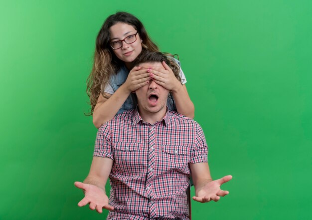 Foto grátis casal jovem, homem e mulher com roupas casuais, mulher feliz, cobrindo os olhos do namorado, fazendo uma surpresa em pé sobre a parede verde