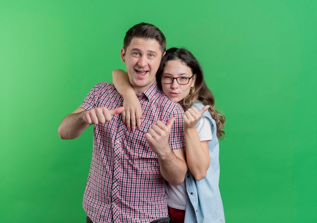 Casal jovem, homem e mulher com roupas casuais, sorrindo, mostrando os polegares em pé sobre a parede verde