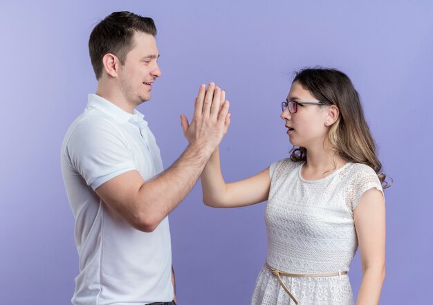 Casal jovem, homem e mulher, olhando um para o outro dando mais cinco em pé sobre a parede azul