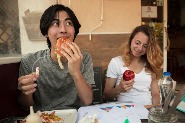 Foto grátis casal jovem multirracial comendo fast food perto de mesa com diagramas