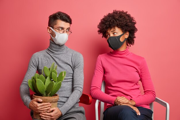 Casal jovem olha tristemente um para o outro usa máscaras faciais durante a quarentena em casa pose em cadeiras para evitar o contágio e fique seguro carregue cacto em vaso