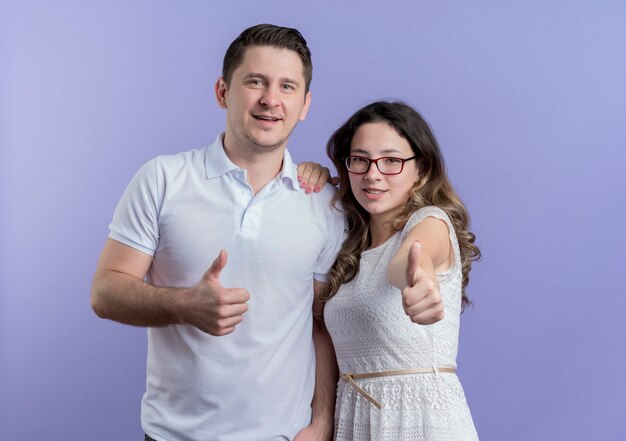 Casal jovem olhando para a câmera sorrindo e mostrando os polegares em pé sobre a parede azul