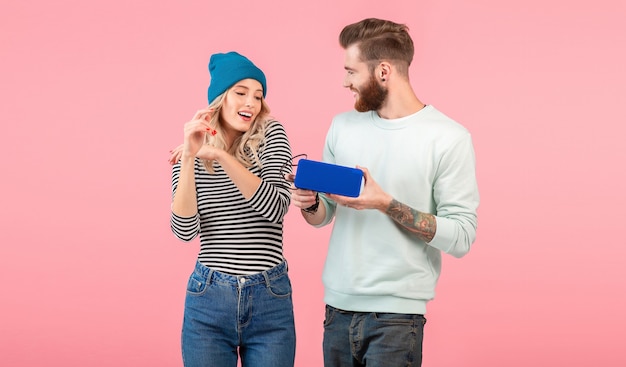 Casal jovem ouvindo música no alto-falante sem fio, vestindo roupas elegantes e legais, sorrindo, posando em rosa