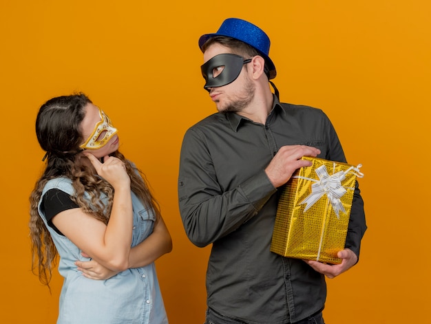 Casal jovem se olha usando máscara de máscara, cara segurando uma caixa de presente isolada em laranja