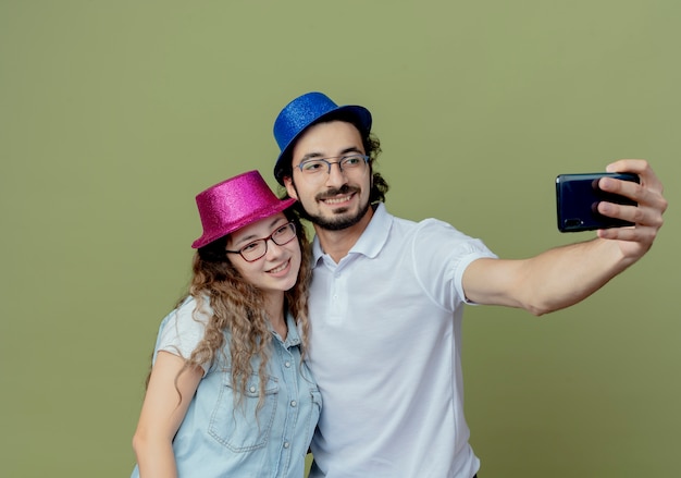 Casal jovem sorridente com chapéu rosa e azul tira uma selfie isolada em verde oliva