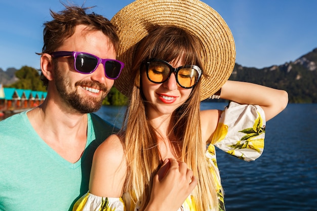 casal marcando selfie perto da incrível vista do lago e montanhas, vestindo acessórios e roupas elegantes. Atmosfera alegre lúdica.