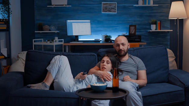 Foto grátis casal relaxado em casa, sentado no sofá, assistindo programas de tv e comendo pipoca, bebendo cerveja, trocando de canal, aproveitando o tempo juntos no lazer em casa, na felicidade e no conceito de pessoas casadas.