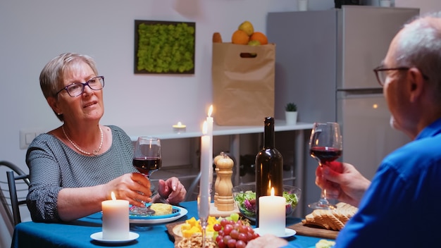 Casal relaxado sênior jantando e bebendo copos de vinho tinto juntos na cozinha em casa. Idosos, idosos aposentados apreciando a refeição, comemorando seu aniversário na sala de jantar.