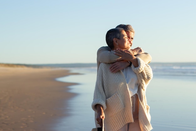 Casal românico sênior passando tempo à beira-mar ao pôr do sol, apreciando a vista fantástica do mar. homem grisalho abraçando sua esposa em pé atrás. romance, aposentadoria, conceito de férias