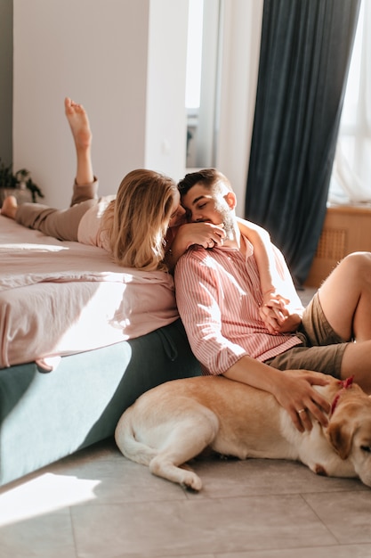 Casal romântico abraços. Homem acaricia labrador e conversa com sua amada deitada na cama.