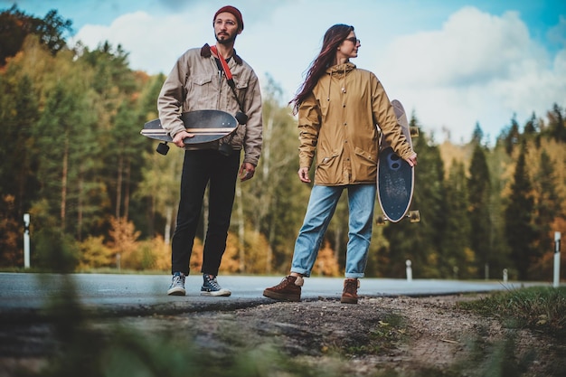 Casal romântico atraente está andando na estrada cercada de árvores de outono enquanto segura seus longboards.