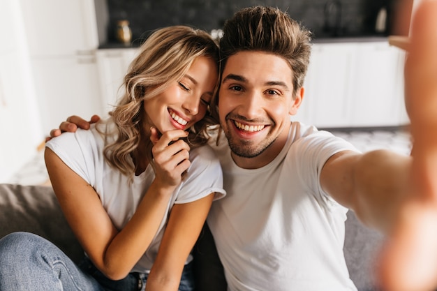 Casal romântico sorridente fazendo selfie em casa sentado no sofá. Homem e namorada sorrindo alegremente com os olhos fechados.