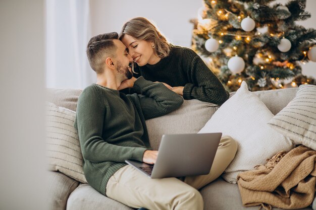 Casal se beijando ao lado da árvore de natal, usando laptop