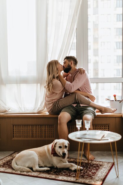 Casal se senta no parapeito da janela e se beija. Menina com roupa bege abraçando o cara enquanto seu labrador está deitado no tapete.