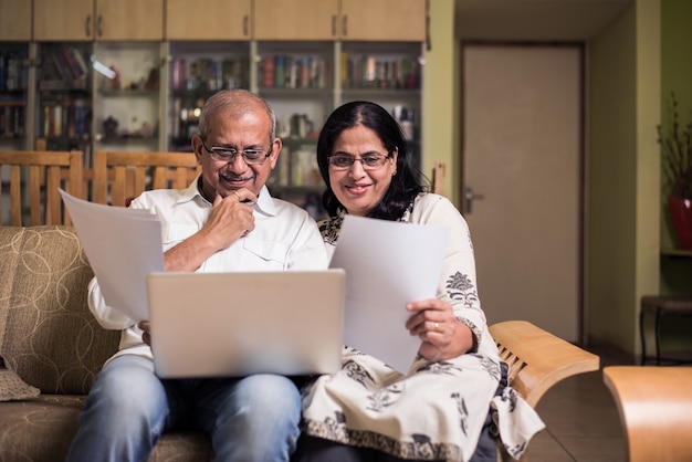 Casal sênior indiano asiático contando, fazendo finanças domésticas e verificando contas com laptop, calculadora e dinheiro também com o cofrinho, enquanto está sentado no sofá, sofá ou mesa em casa