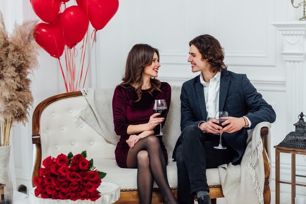 Casal sorridente, sentado no sofá, bebendo vinho e olhando um para o outro