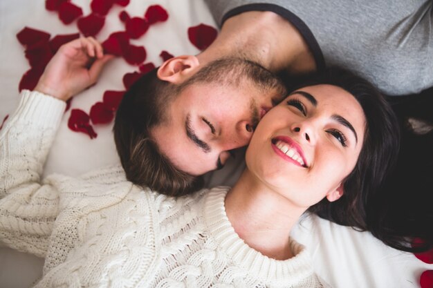 Casal sorrindo e mentindo sobre a cabeceira da cama com a cabeça rodeada de pétalas de rosa