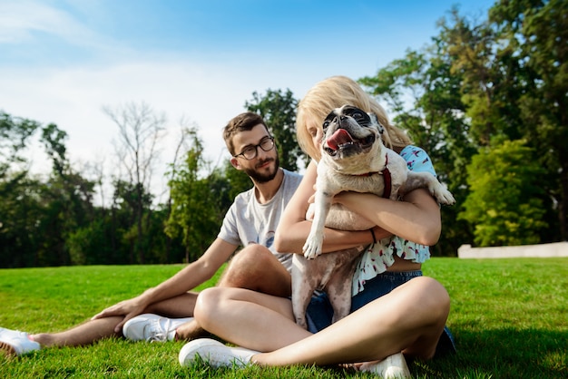 Casal sorrindo, sentado na grama com bulldog francês no parque