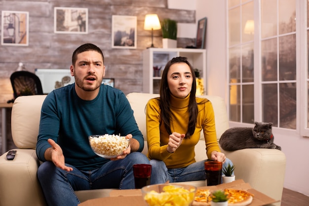 Casal torcendo pelo time favorito enquanto assiste TV e come fast food