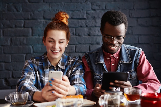 Casal usando aparelhos modernos enquanto relaxa no café. mulher ruiva lendo informações na página da web via telefone celular enquanto homem negro assistindo vídeo no tablet digital
