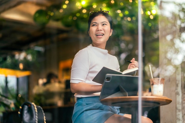 Casual relaxa mulher asiática freelance nômade digital escrevendo idéias de caderno de desenho notadas por seu novo projeto no café, filmado pela janela da cafeteria mulher asiática sentada trabalhando no café sentimento alegre