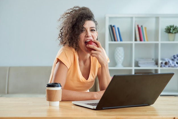 Cauasian jovem encaracolado comendo maçã na frente do laptop em casa