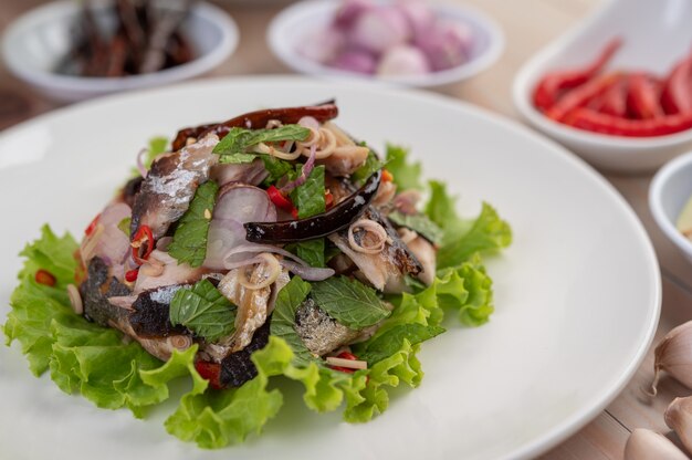 Cavala frita coberta com galanga, pimenta, hortelã, cebola vermelha em um prato branco.