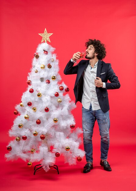 Celebração de natal com um jovem barbudo bebendo seu vinho em pé perto da árvore de natal no vermelho
