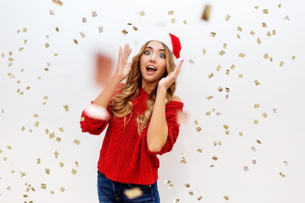 Celebrando a garota com chapéu de baile de máscaras de papai noel se divertindo em confetes na parede branca. novo clima de festa na orelha. pulôver vermelho aconchegante. emoções verdadeiras. surpreenda emoções malucas.