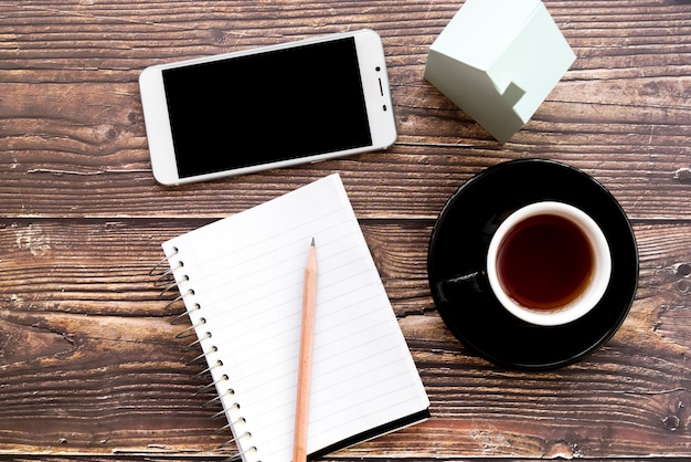 Celular; caderno espiral em branco; lápis; xícara de café e casa modelo na mesa de madeira