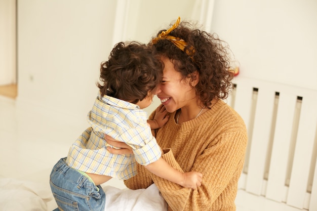 Cena aconchegante de uma jovem mãe de cabelos cacheados feliz e feliz, acariciando o filho bebê em seus braços, unindo-se no quarto, desfrutando da maternidade, sentindo uma profunda conexão com seu filho pequeno. Amor e felicidade