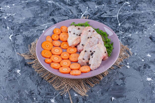 Cenouras fatiadas, asas e verduras em um prato sobre uma serapilheira na superfície azul