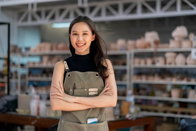 Ceramista feminina da Ásia com peito confiante de mão de avental em seu estúdio de escultura em argila de oficina com boas-vindas calorosas e sorridentes positivas prontas para iniciar uma nova oficina de cerâmica de fábrica com nova coleção de trabalho