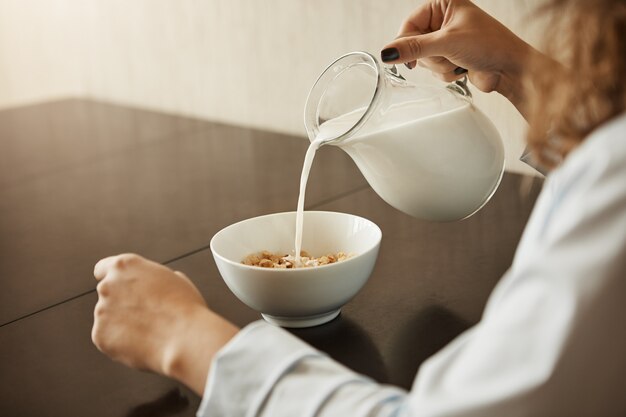 Cereais é o melhor café da manhã para manter a forma. Foto recortada de mulher sentada em roupa de noite, derramando leite na tigela com cereais, preparando a refeição para comer e correu para o trabalho, ouvindo notícias na TV