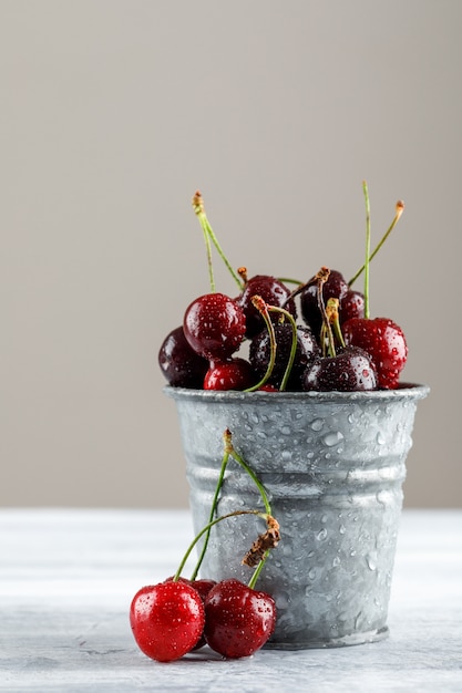 Cerejas em um mini balde no grunge e superfície cinza, vista lateral.